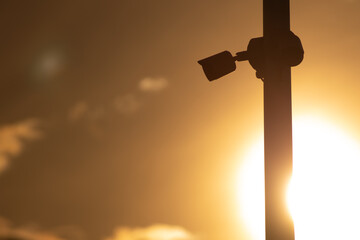 A silhouetted security camera mounted on a pole is captured against a vibrant sunset, with the sun glowing brightly in the background. The image emphasizes the contrast between technology and nature