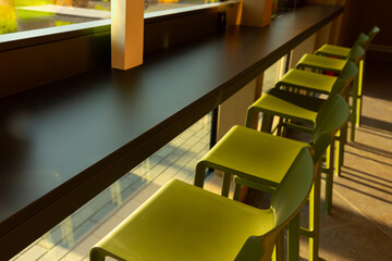 A row of sleek green chairs lined up along a black bar counter in a modern interior. Sunlight streams in through large windows, casting warm, inviting shadows on the floor and enhancing the vibrant
