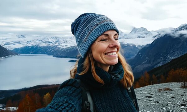 Full-length portrait video of a grinning woman in her 40s wearing a cool cap or hat against a nordic or scandinavian landscape background