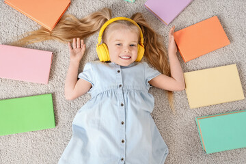 Cute little girl in headphones listening to audiobook with books while lying on carpet at home