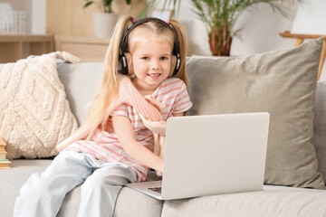 Cute little girl in headphones listening to audiobook with modern laptop while sitting on sofa in living room