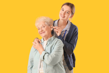 Nurse putting hearing aid on senior woman against yellow background