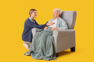 Nurse with stethoscope listening to senior woman in armchair on yellow background