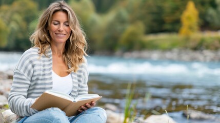 Obraz premium A woman enjoys reading a book while sitting by the riverside in a peaceful natural setting, surrounded by trees and flowing water, symbolizing calm and relaxation.
