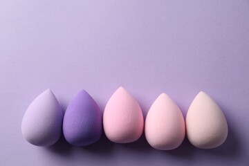 Many colorful makeup sponges on violet background, flat lay. Space for text