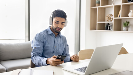 Serious young Arab student man in headphones using smartphone ,laptop for online learning at home, typing on mobile phone, sitting at workplace table. Employee listening music while working