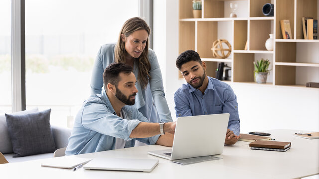 Three young multiethnic business colleagues watching website presentation on pc, sitting at workplace. Project manager showing online content to coworkers, pointing hand at laptop screen