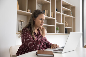 Focused mid adult business professional woman in casual working at laptop at workplace table, touching chin, thinking on career, modern technology for job, reading online content