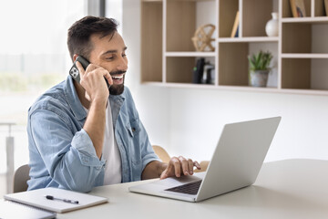 Cheerful handsome Hispanic entrepreneur man talking on cellphone at workplace, typing on laptop,...