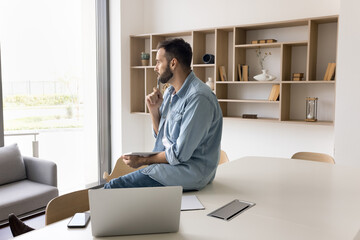 Serious thoughtful handsome entrepreneur man in casual sitting at table, leaning at large workplace desk, looking at window away in deep thoughts, thinking pn project reports, using digital gadgets