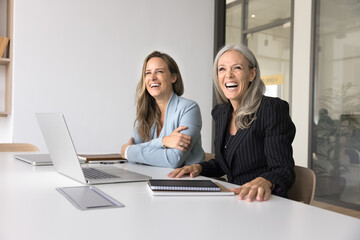 Cheerful younger and older female business colleagues sitting at workplace table in office, looking away, smiling, laughing, working at laptop together, promoting professional communication