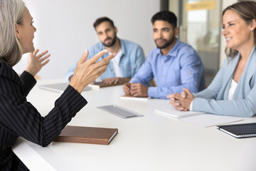 Confident older business leader woman talking to diverse team of younger colleagues at meeting, sitting at large table, giving seminar, training, coaching. Female boss speaking to co-workers