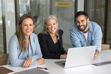 Successful team of three younger and elder business colleagues sitting at workplace table in office, looking at camera, smiling for portrait, promoting professional communication, working together