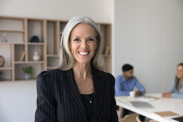 Happy female senior business leader woman posing for office portrait, standing in meeting room with colleagues working and talking in background, looking at camera, smiling for head shot