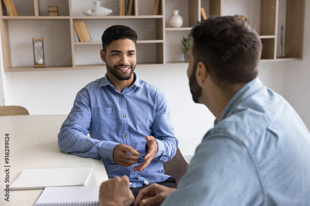 Wall mural positive handsome young arab professional man talking to male colleague at workplace table, speaking