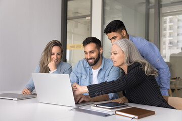 Successful diverse group of business colleagues focused on laptop watching online content, project presentation, pointing at screen, smiling, laughing, sitting at office meeting table