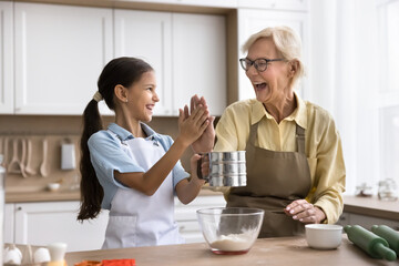 Excited grandma and happy grandkid girl cooking pastry in home kitchen together, sifting flour into...
