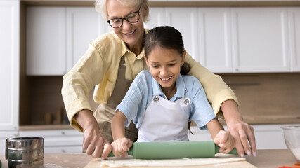 Obraz premium Joyful blonde granny helping cute grandkid girl to roll dough in home kitchen. Happy grandma and granddaughter kid enjoying family culinary baking hobby, preparing bakery food. Banner shot
