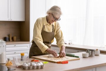 Positive elder retired baker lady cooking biscuits in home kitchen, cutting fresh dough for star shaped cookies, smiling, baking sweet snacks, bakery dessert for at table with ingredients
