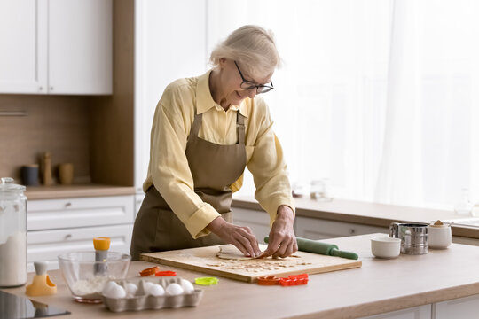 Positive elder retired baker lady cooking biscuits in home kitchen, cutting fresh dough for star shaped cookies, smiling, baking sweet snacks, bakery dessert for at table with ingredients