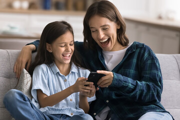 Cheerful young mom and happy cute daughter kid using application on smartphone for online communication at home, watching video on Internet, having fun, shouting for joy