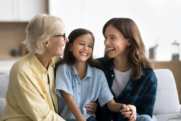 Joyful young mom and senior grandma cuddling kid girl, tickling child, laughing, giggling, enjoying family leisure, close relationship. Happy girls and women of three generations having fun at home
