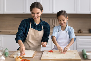 Happy mom and cute kid girl wearing kitchen aprons, baking cookies together, cutting raw dough with molds, smiling, laughing, enjoying family culinary hobby, preparing sweet dessert, bakery food