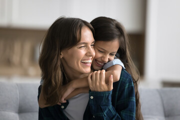 Cheerful loving little kid girl hugging positive mom from behind, playing with mummy at home. Happy mother piggybacking sweet child, playing active family game, laughing