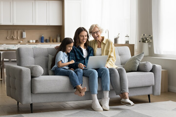 Obraz premium Happy blonde older grandma, young mom and cute kid girl using laptop on home couch, watching movie online, talking on video call, using computer for internet communication in cozy apartment