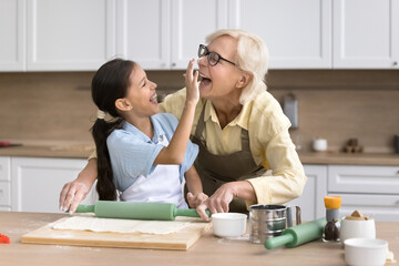 Cheerful grandchild girl and grandma baking and having fun in home kitchen, cooking bakery food together. Happy kid touching grandmothers face with floury finger, laughing