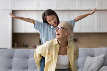 Happy excited strong grandma piggybacking granddaughter, sitting on home sofa, enjoying healthy back, laughing. Cheerful kid playing airplane, riding granny back, enjoying family game