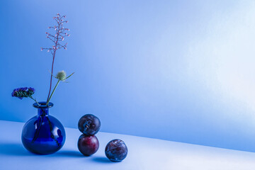Still life with ripe plums, blue glass vase with a bouquet of flowers on a blue background....
