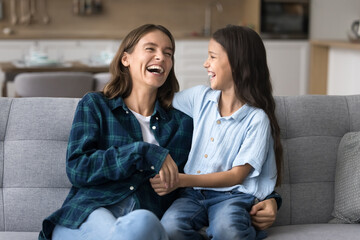 Joyful excited mom and sweet little kid girl hugging and having fun on home couch, laughing at...