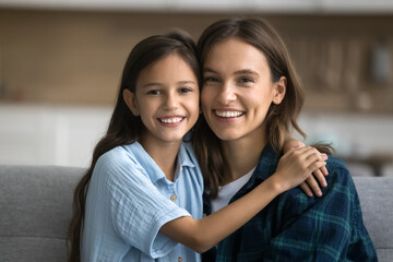 Cheerful beautiful mom and happy daughter child looking at camera, hugging with head touches, smiling, posing for home portrait. Positive mother and cute girl head shot