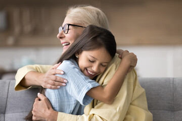 Happy adorable grandkid girl tightening beloved granny with closed eyes at home. Grandma and child...