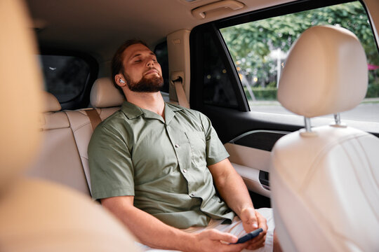 A man relaxing in the back seat of a car, wearing wireless earphones and holding a smartphone, exuding a calm and comfortable atmosphere