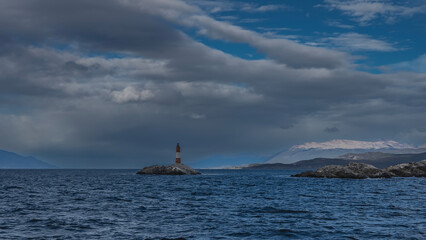 Fototapeta premium An old lighthouse on a tiny rocky island in the ocean. A red and white striped tower against the sky, clouds. Mountains in the distance. Ripples on the surface of the blue water of the Beagle Channel