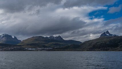 The picturesque mountain range of the Andes. Snow-capped peaks against a blue sky, clouds. The city houses of Ushuaia are at the foot, on the ocean shore. The surface of the Beagle Canal. Argentina.