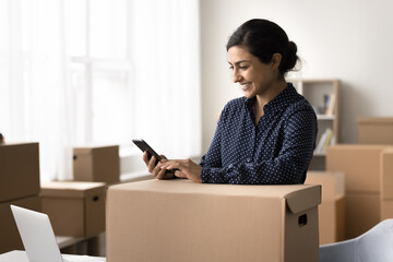 Smiling Indian woman hold smartphone working online in warehouse, get notification about incoming order or parcel that need to be prepared for dispatch, checking order details using mobile application