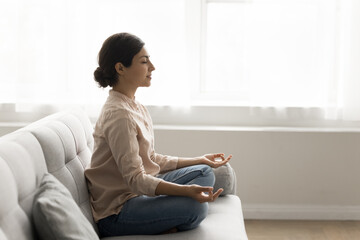 30s woman sit on sofa meditating at home, calming mind, relax body, reducing stress levels,...