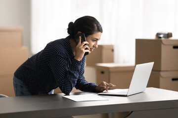 Woman talk on phone, check orders use laptop working in warehouse, ordering delivery services, makes calls to supplier, communicate with colleagues, coordinate workflow tasks or address urgent matters