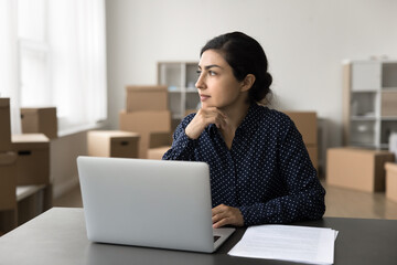 Pensive Indian woman sit at desk with laptop, thinking on marketing campaign development or ideas, working in warehouse, heap of cardboard boxes on background. Drop shipping, electronic commerce, SEO