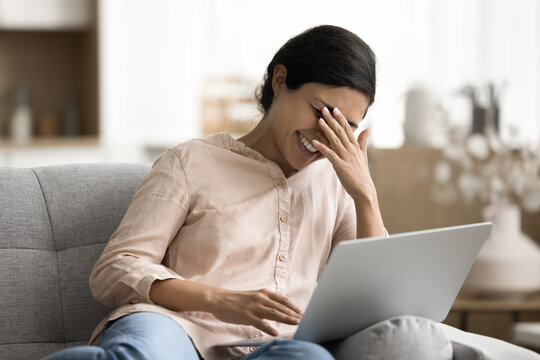 Cheerful young Indian woman relaxing on couch with wireless computer, laughing while watching on-line movie using digital subscription streaming services, enjoy funny tv show or video. Leisure, fun