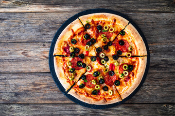 Circle pizza chorizo with black and green olives slices and mozzarella served on black stony plate on wooden table
