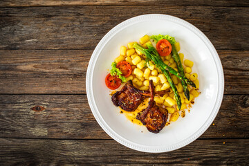 Lamb chops with bone, gnocchi and cooked green asparagus served on white plate on wooden table
