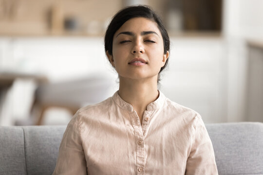 Young serene Indian woman meditates with eyes closed to boost state of inner peace, mindfulness and self-awareness, female enjoy moment of tranquillity, relaxing, focusing on her thoughts, quiets mind