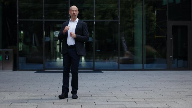 A businessman dressed in dark jacket and white shirt is dancing near glass office building. Man is walking with joy, in good mood. He is celebrating completed plan, successful deal. Full shot