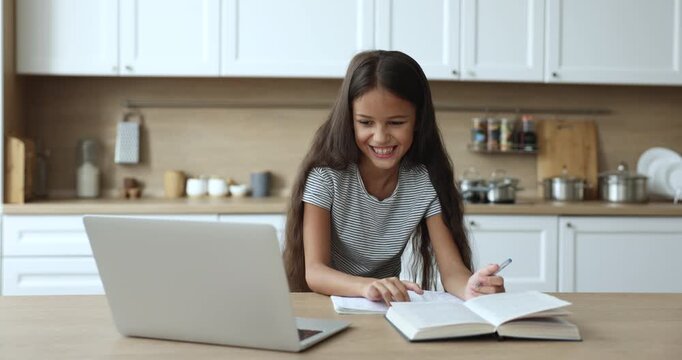 Positive smart early schoolkid girl studying online at home, talking to teacher on video call an laptop, writing learning notes in copybook, attending virtual class on Internet, smiling