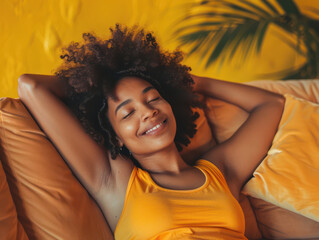 A young African woman is relaxing on a couch, smiling and looking peaceful. She is wearing a yellow tank top and has curly hair.