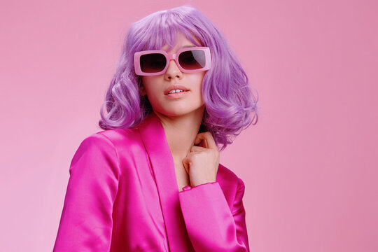 Purplehaired woman in sunglasses and a pink suit, posing confidently against a matching pink background, exuding a bold and stylish fashion statement with her unique and vibrant appearance